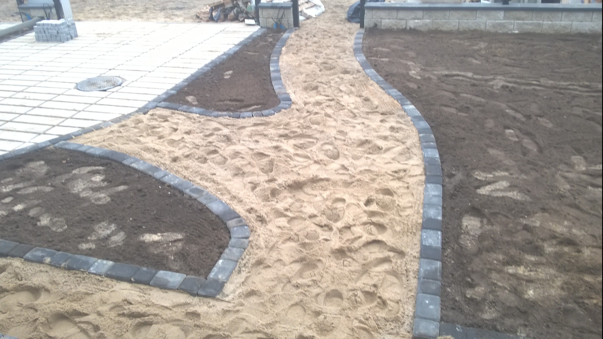 Kształtowana ścieżka z piasku i obrzeży z kostki brukowej, fragment trawnika z świeżo nawiezioną ziemią, w tle ułożona kostka brukowa.