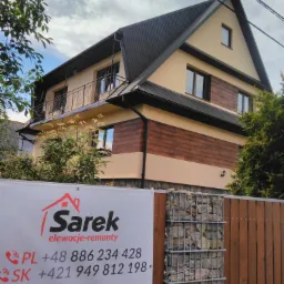 Dwukondygnacyjny dom z beżową elewacją i elementami dekoracyjnymi imitującymi drewno, widoczny zza ogrodzenia z gabionami i baneru reklamowego firmy Sarek oferującej elewacje i remonty.