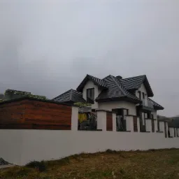 Nowoczesny dom z ciemną dachówką i białą elewacją, otoczony murem z drewnianymi elementami, widoczne materiały budowlane na dachu, pochmurne niebo.