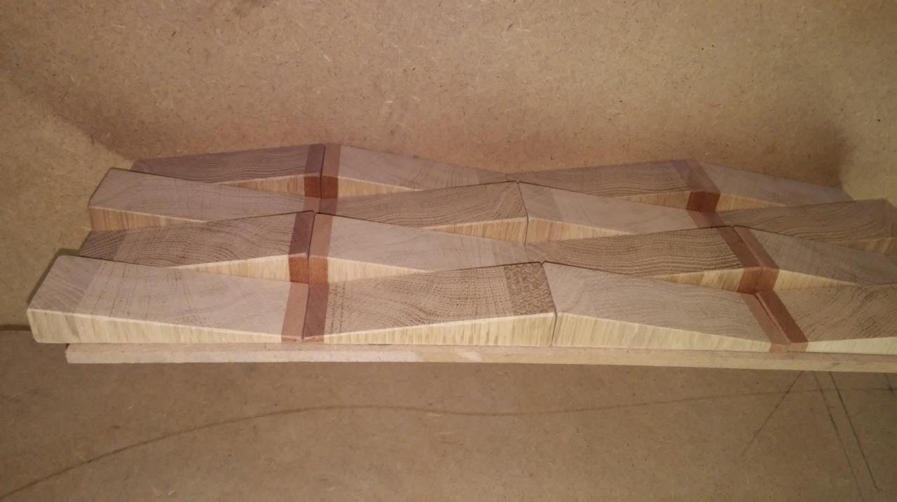 Surowe, drewniane elementy dekoracyjne z ukośnymi krawędziami i wstawkami z ciemniejszego drewna, ułożone na płycie, prawdopodobnie w trakcie montażu.