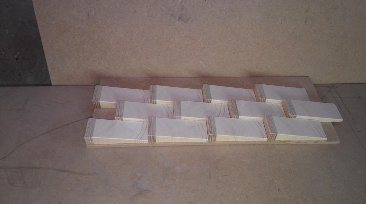 Przygotowane drewniane elementy trapezowe z widocznymi warstwami sklejki, ułożone na płycie wiórowej, prawdopodobnie do dalszej obróbki lub montażu.