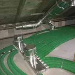 Zielone rury wentylacyjne ułożone równolegle na poddaszu, z elementami srebrnej izolacji i widoczną konstrukcją dachu z drewnianych belek