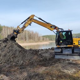KANKIEWICZ ROBOTY ZIEMNE sp. z o.o. - Żółta koparka CAT M313D nabiera ziemię z hałdy na tle jeziora i lasu. Operator w kabinie. Widoczne ślady opon. Błękitne niebo.