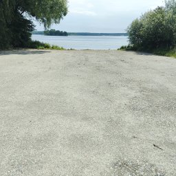 KANKIEWICZ ROBOTY ZIEMNE sp. z o.o. - Betonowy zjazd do jeziora otoczony zielenią, w oddali widoczny ląd. Jasne niebo i spokojna woda tworzą malowniczy krajobraz. Widok z perspektywy zjazdu.