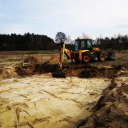 Żółta koparko-ładowarka JCB w trakcie prac ziemnych na budowie, widoczne wykopane fundamenty i hałdy ziemi, w tle las i błękitne niebo.