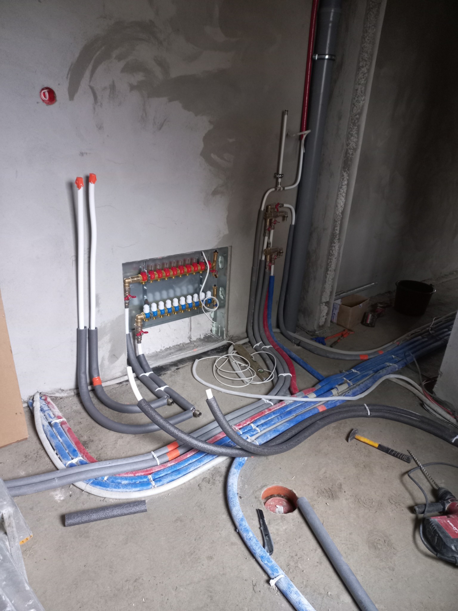 Rozdzielacz ogrzewania podłogowego z rurami w izolacji, w trakcie montażu w surowym wnętrzu, widoczne narzędzia i instalacje.