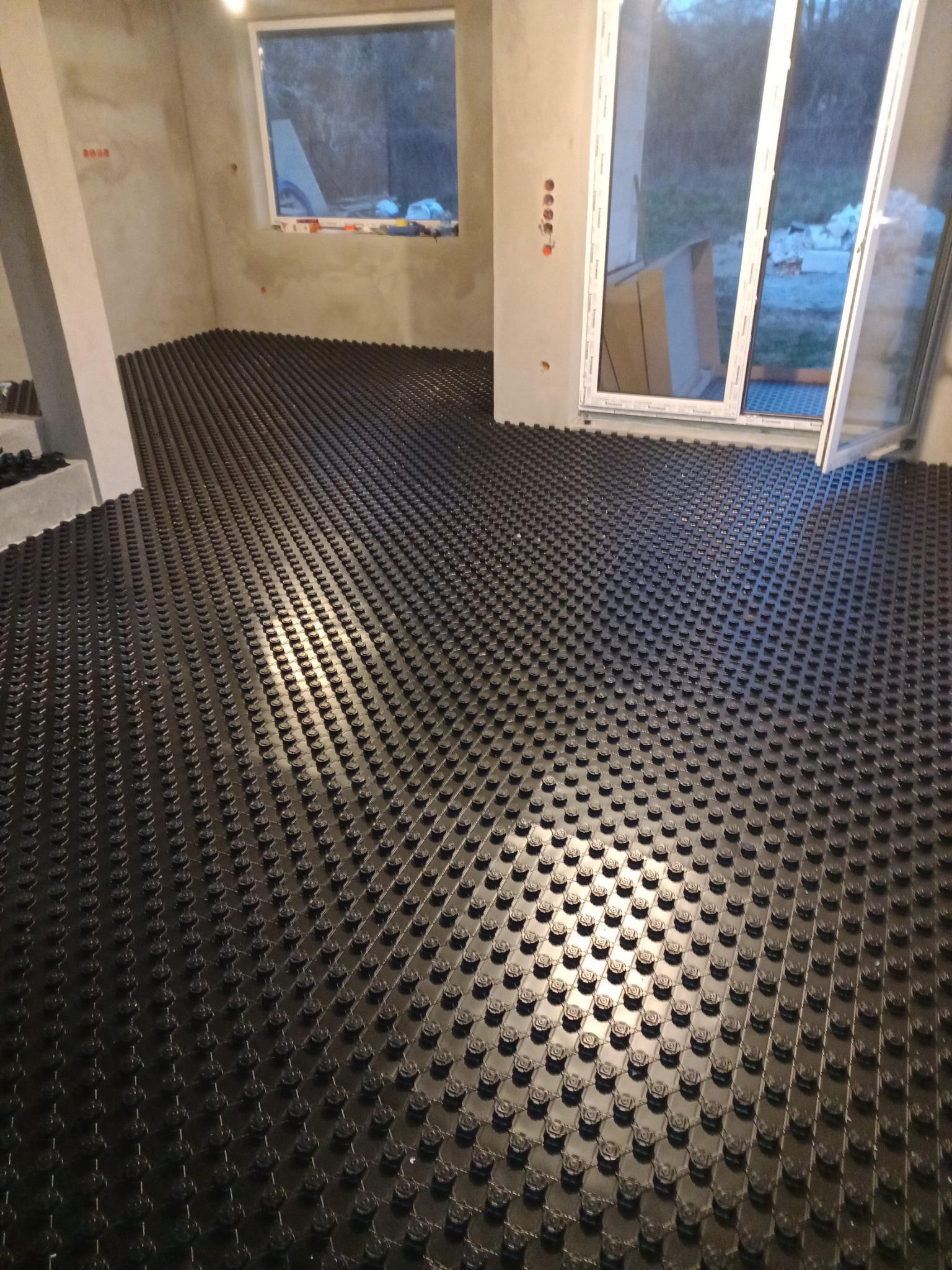 Czarne, plastikowe maty grzewcze pod podłogówkę, rozłożone równomiernie w surowym pomieszczeniu z oknem i otwartymi drzwiami balkonowymi, widoczne niedoskonałości ścian.