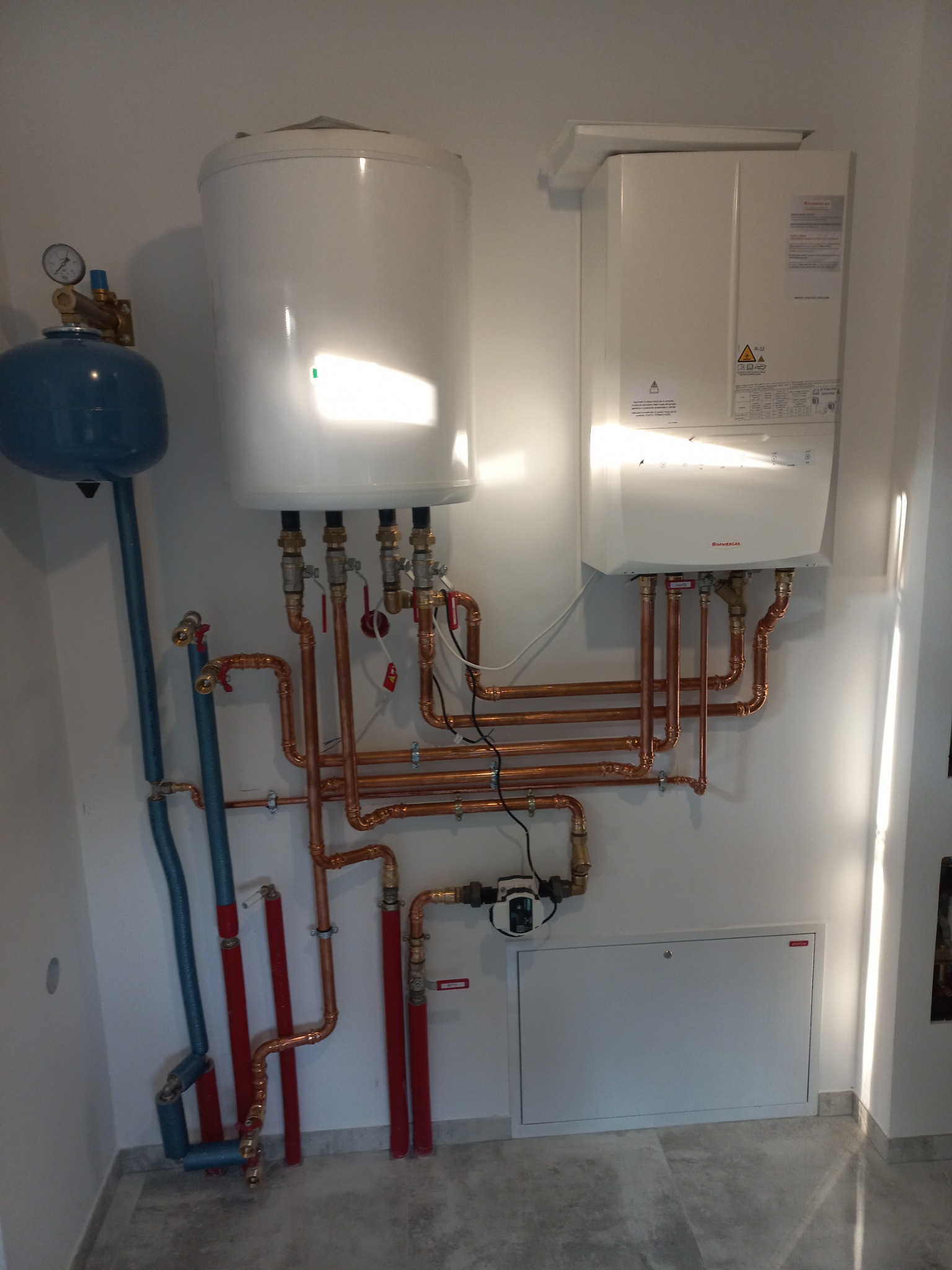 Biały zasobnik ciepłej wody i piec gazowy wiszące na ścianie z widoczną instalacją hydrauliczną z rur miedzianych i niebieskich, oraz szarym tłem podłogi.