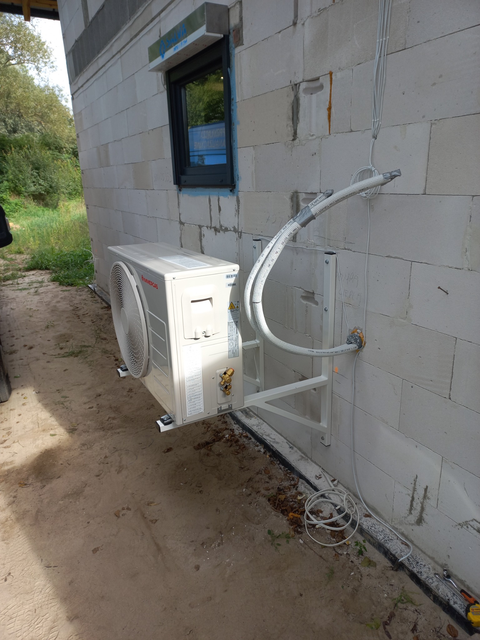 Zewnętrzna jednostka klimatyzacji Immergas zamontowana na białej ścianie z betonu komórkowego. Widoczne przewody w izolacji, mocowanie na metalowym wsporniku, okno w tle i ziemia pod jednostką.