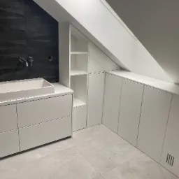 Białe szafki wnękowe pod skosem dachu, z otwartą półką, umywalka nablatowa na szafce z szufladami, czarna ściana z teksturą, szara podłoga