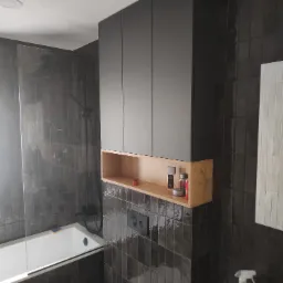 Nowoczesna łazienka z szarymi, błyszczącymi płytkami, białą wanną z parawanem, toaletą i wiszącą szafką z czarnymi frontami i otwartą półką z jasnego drewna, na której stoją kosmetyki.