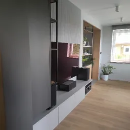 Nowoczesna zabudowa ściany telewizyjnej z szarymi szafkami, półkami z drewna i metalu oraz dekoracyjną ścianką z pionowymi żłobieniami, uzupełniona o głośniki i rośliny doniczkowe.