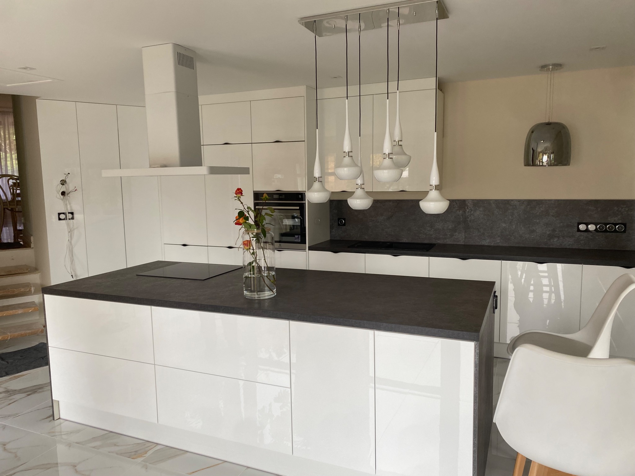Nowoczesna kuchnia z białymi, lakierowanymi frontami, wyspą z czarnym blatem i szklanym wazonem z kwiatami na blacie, wiszące lampy nad wyspą.