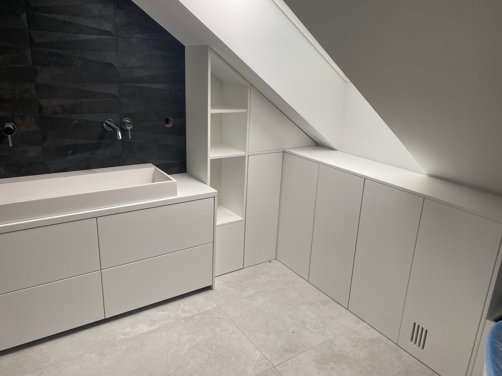 Białe szafki wnękowe pod skosem dachu, z otwartą półką, umywalka nablatowa na szafce z szufladami, czarna ściana z teksturą, szara podłoga