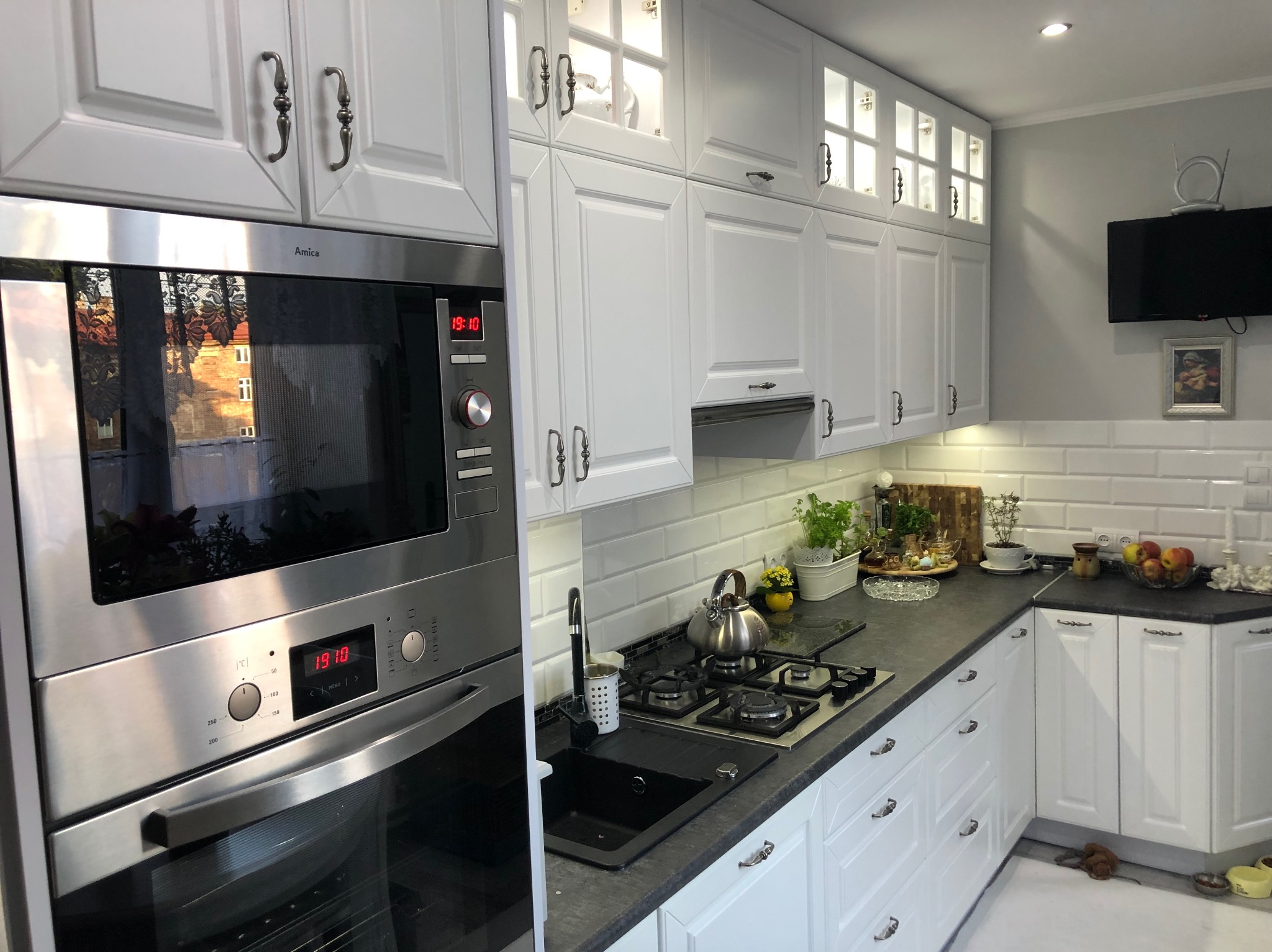 Biała kuchnia z zabudową, fronty z ozdobnymi uchwytami, piekarnik Amica z odbiciem budynku w szybie, szary blat, białe płytki na ścianie i telewizor.