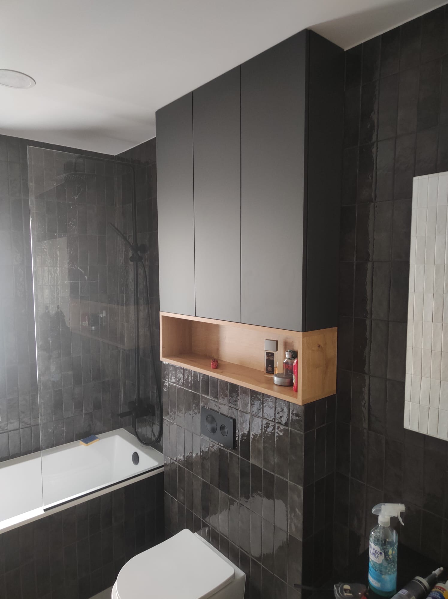 Nowoczesna łazienka z szarymi, błyszczącymi płytkami, białą wanną z parawanem, toaletą i wiszącą szafką z czarnymi frontami i otwartą półką z jasnego drewna, na której stoją kosmetyki.