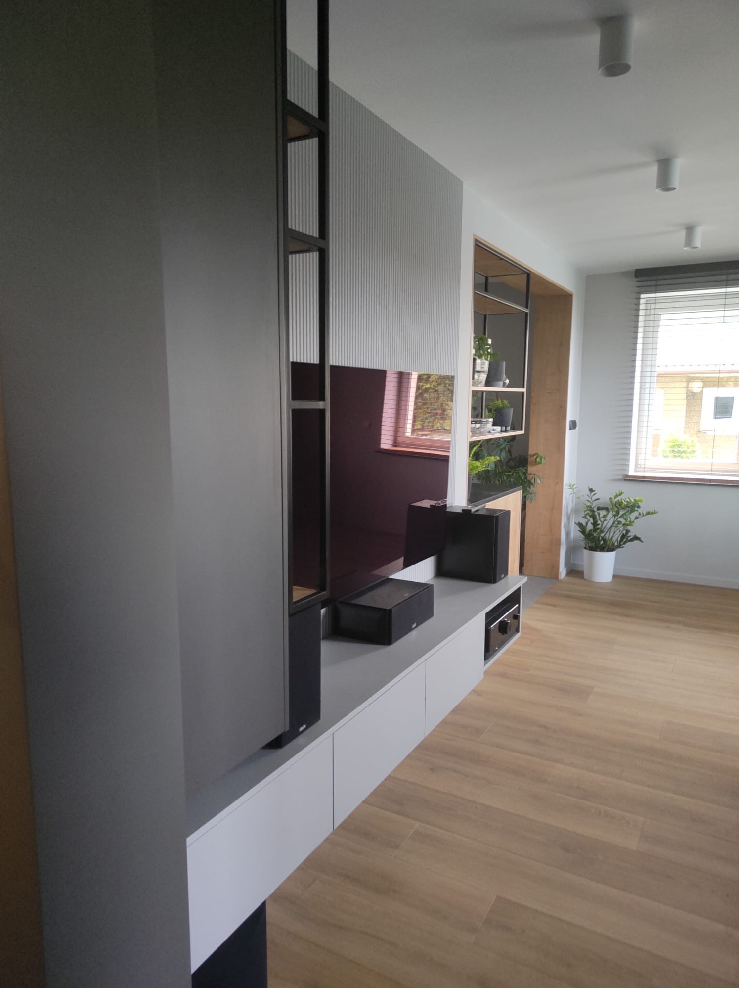Nowoczesna zabudowa ściany telewizyjnej z szarymi szafkami, półkami z drewna i metalu oraz dekoracyjną ścianką z pionowymi żłobieniami, uzupełniona o głośniki i rośliny doniczkowe.