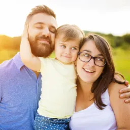 Uśmiechnięta rodzina: ojciec z brodą, matka w okularach i mała dziewczynka przytuleni na tle zielonej łąki w słoneczny dzień.