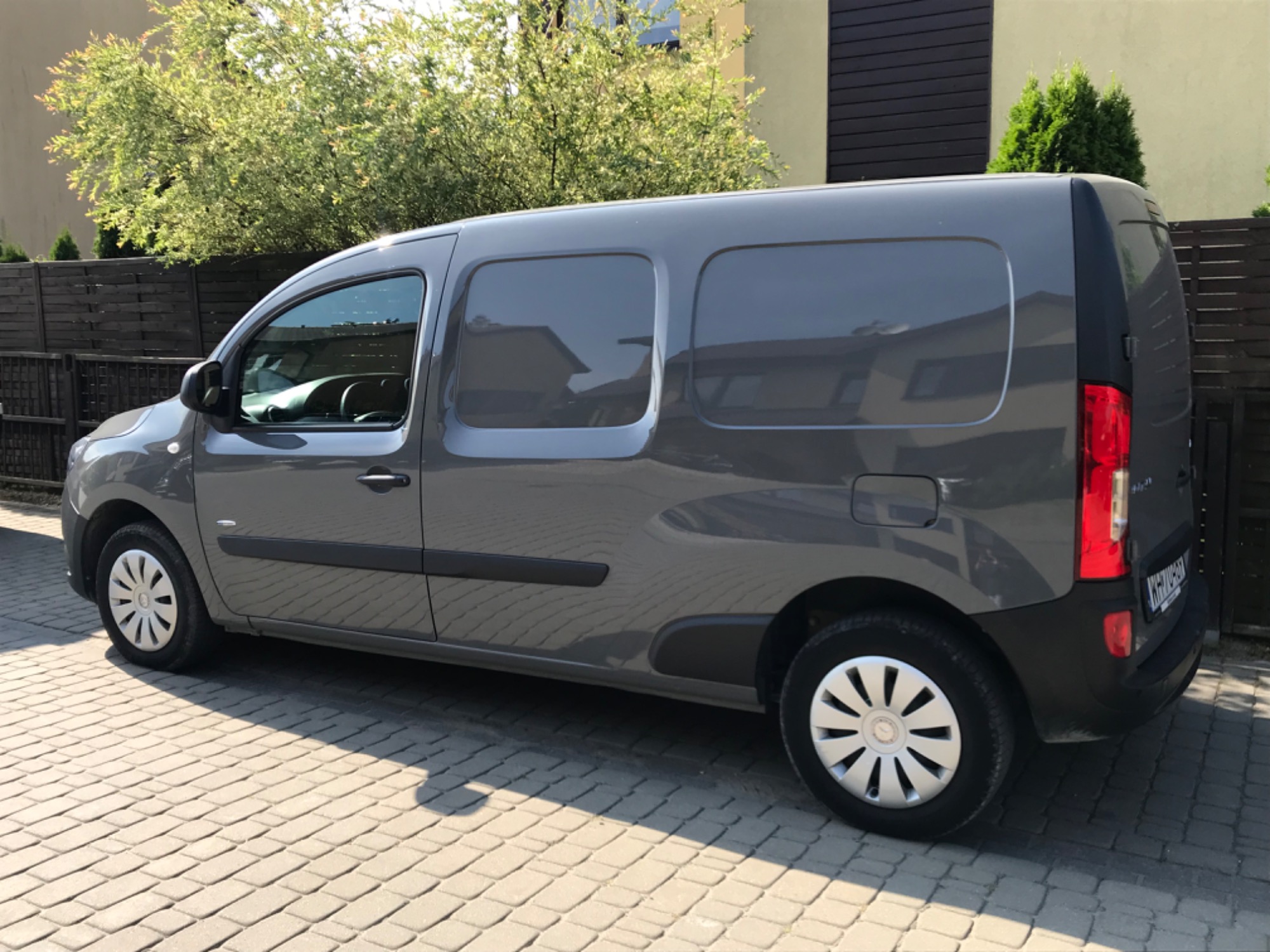 Szary bus Renault Kangoo z zaciemnionymi szybami, zaparkowany na kostce brukowej przed budynkiem z zielenią w tle, widok z boku.