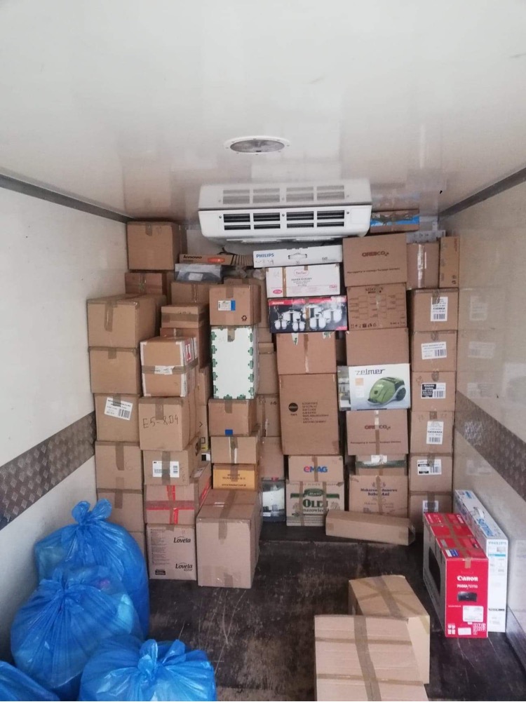 Wnętrze chłodni wypełnione kartonami z różnymi przedmiotami, zabezpieczonymi niebieskimi workami foliowymi, widoczna klimatyzacja na suficie.