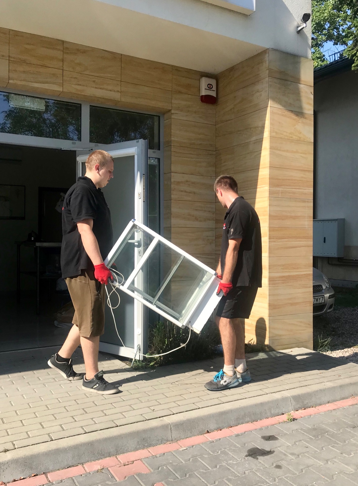 Dwóch mężczyzn w czerwonych rękawicach przenosi białą ramę okienną na zewnątrz budynku o elewacji z paneli imitujących drewno.