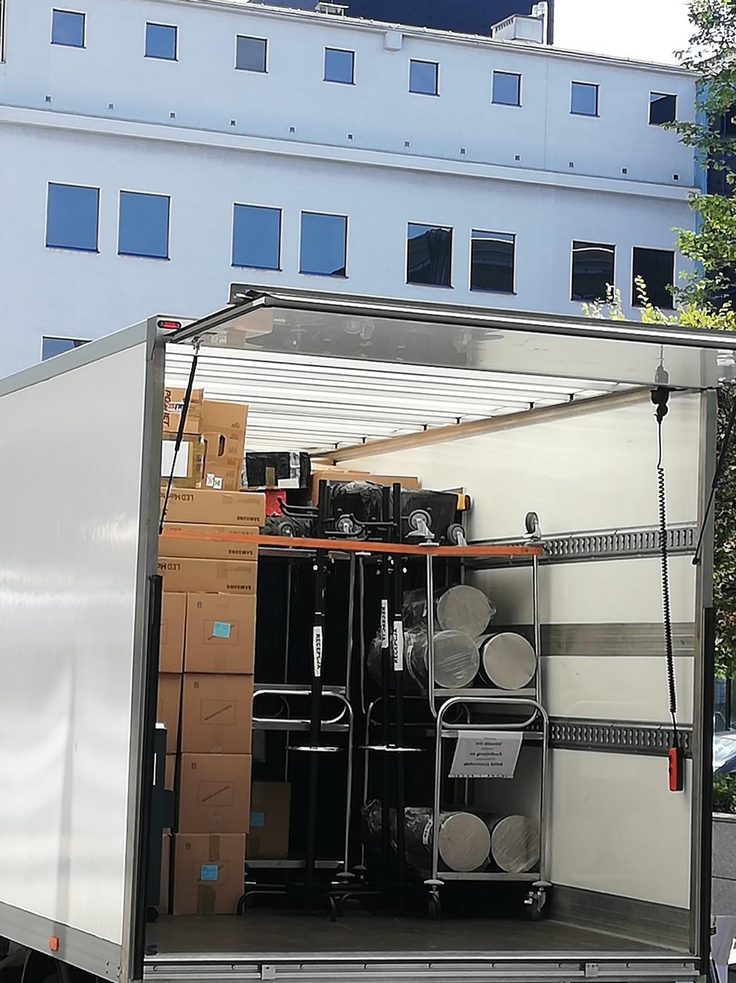 Wnętrze otwartego busa załadowane kartonami z logo Samsung i metalowymi wózkami z walcami, na tle budynku z regularnymi oknami.