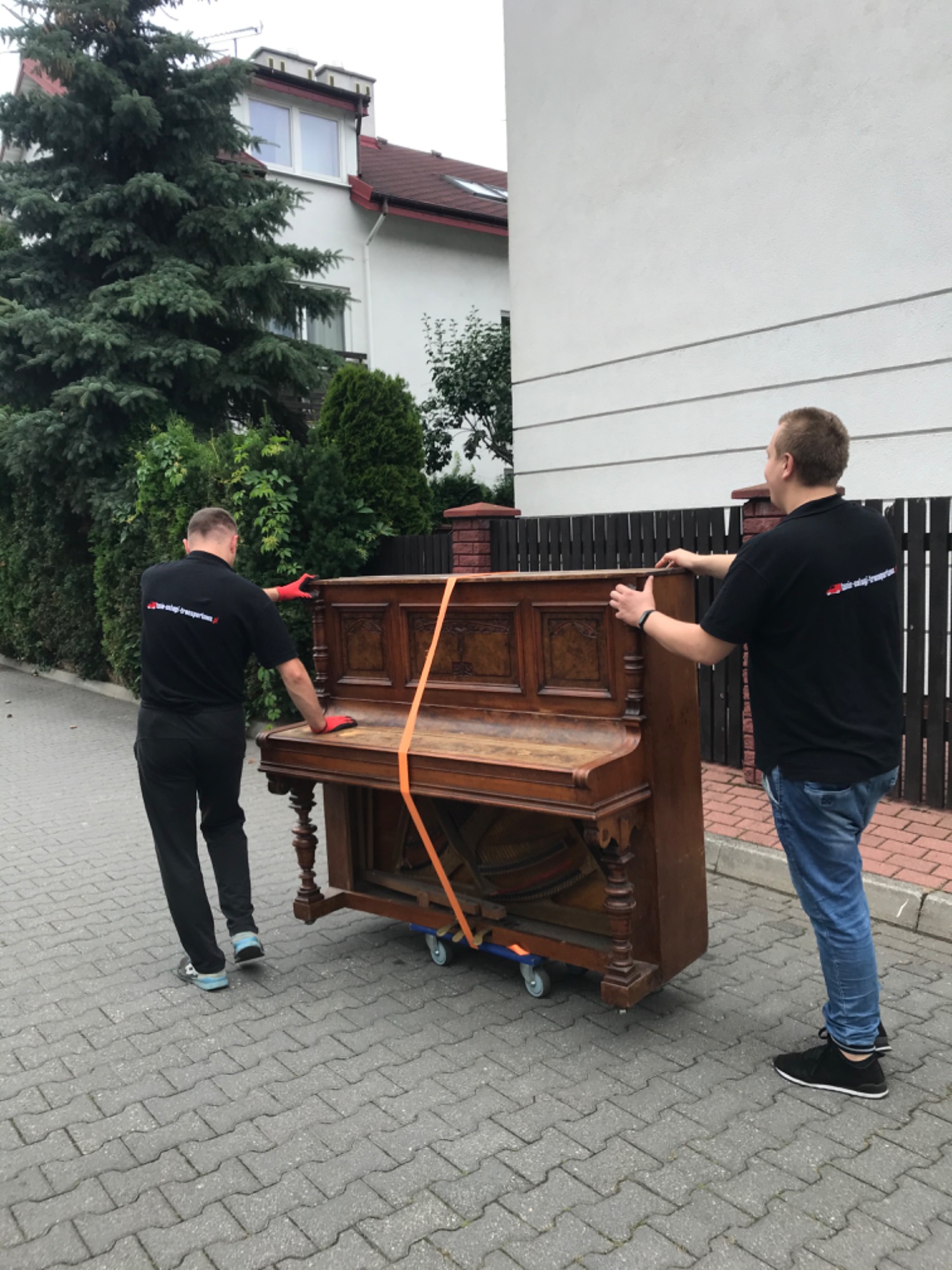 Dwóch mężczyzn w czarnych koszulkach z logo firmy transportowej, w czerwonych rękawicach, przenosi zabytkowe pianino zabezpieczone pomarańczowymi pasami transportowymi na wózku z kółkami, po...