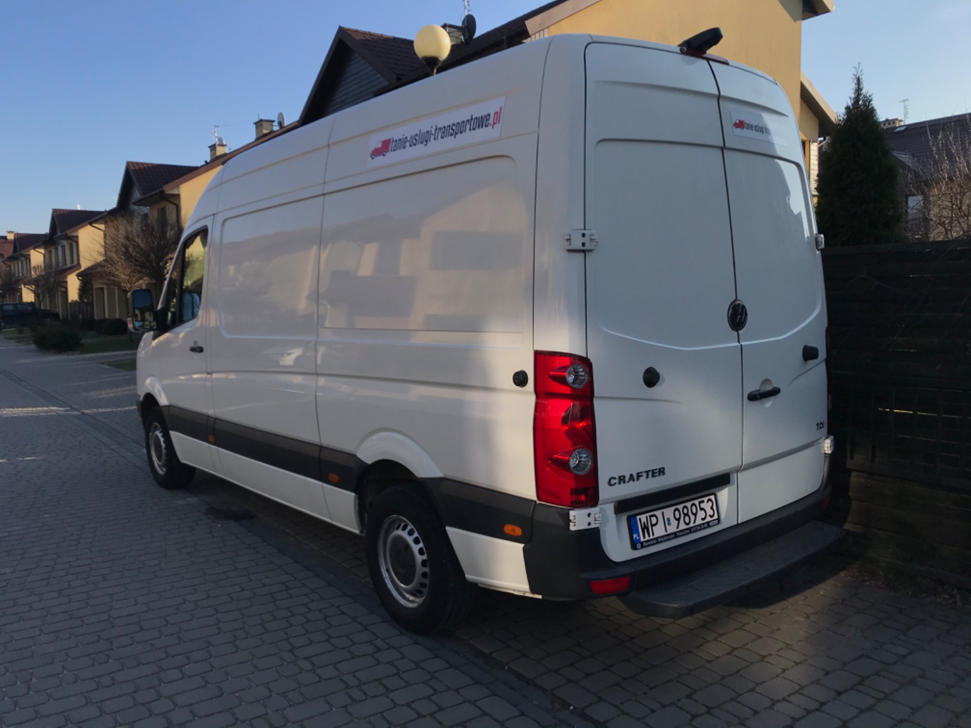 Biały bus marki Volkswagen Crafter zaparkowany na brukowanej ulicy w dzielnicy domów jednorodzinnych, na boku naklejka z adresem strony internetowej.