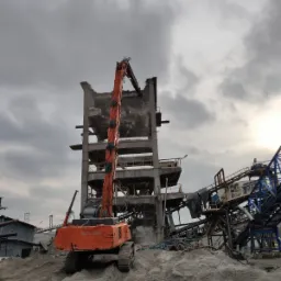 Pomarańczowa koparka Hitachi z długim ramieniem kruszy betonowy szkielet budynku przemysłowego na tle pochmurnego nieba, z widocznymi elementami konstrukcji i gruzem.