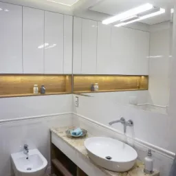 Nowoczesna łazienka z owalną umywalką nablatową na blacie z jasnego kamienia, białe szafki wiszące z podświetlanymi półkami, bidet i drewnopodobna podłoga.