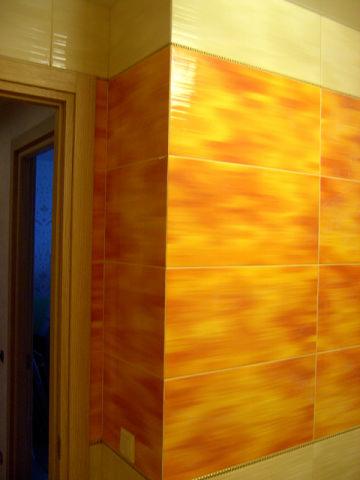 Żółto-pomarańczowe płytki imitujące drewno na ścianie w łazience, widoczny narożnik i fragment ościeżnicy drzwi.