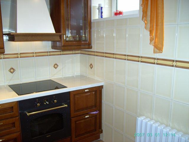 Wykończona kuchnia z beżowymi płytkami z dekoracyjnym pasem, ciemne drewniane szafki z czarnym piekarnikiem i płytą indukcyjną, biały blat i grzejnik.