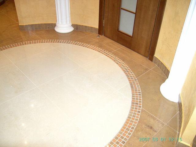 Beżowa, okrągła posadzka z płytek w przedpokoju, obramowana mozaiką, z kolumną i drzwiami w tle.