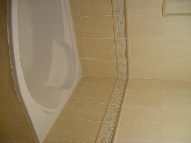 Biała wanna narożna w beżowej łazience, wykończona płytkami z dekoracyjnym pasem.