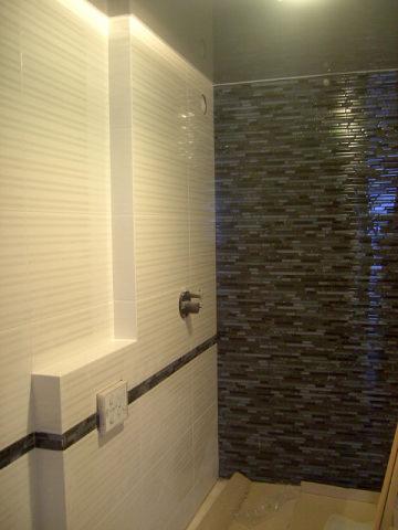 Wykończona łazienka z jasnymi płytkami imitującymi drewno, ciemnym pasem dekoracyjnym i wnęką, oraz ścianą prysznicową wyłożoną mozaiką z kamienia naturalnego.