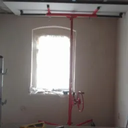 Wnętrze w trakcie remontu: podnośnik do płyt gipsowo-kartonowych z zamontowaną płytą, surowy sufit z widocznymi elementami konstrukcyjnymi, ściany przygotowane pod malowanie, narzędzia na stole...