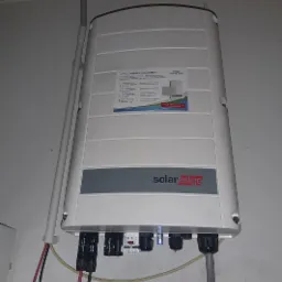 Biała obudowa falownika SolarEdge zamontowana na ścianie, widoczne kable i przewody elektryczne.