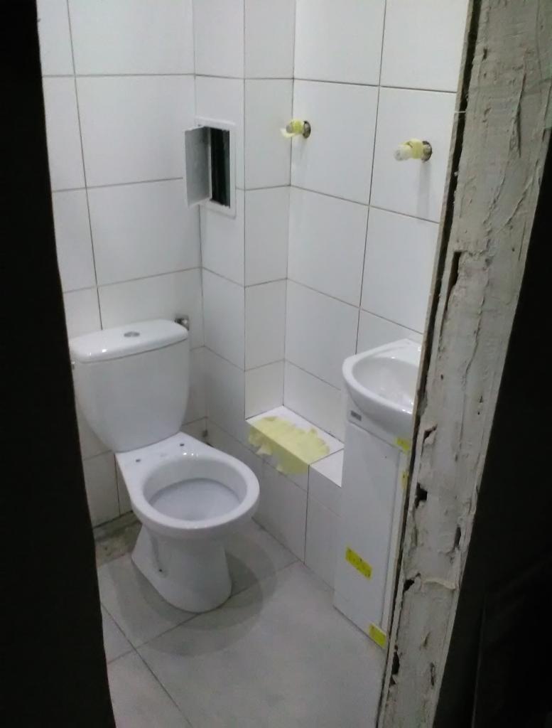 Biała toaleta i umywalka w świeżo wyremontowanej łazience z białymi płytkami, zabezpieczone żółtą taśmą malarską, widok zza framugi drzwi.