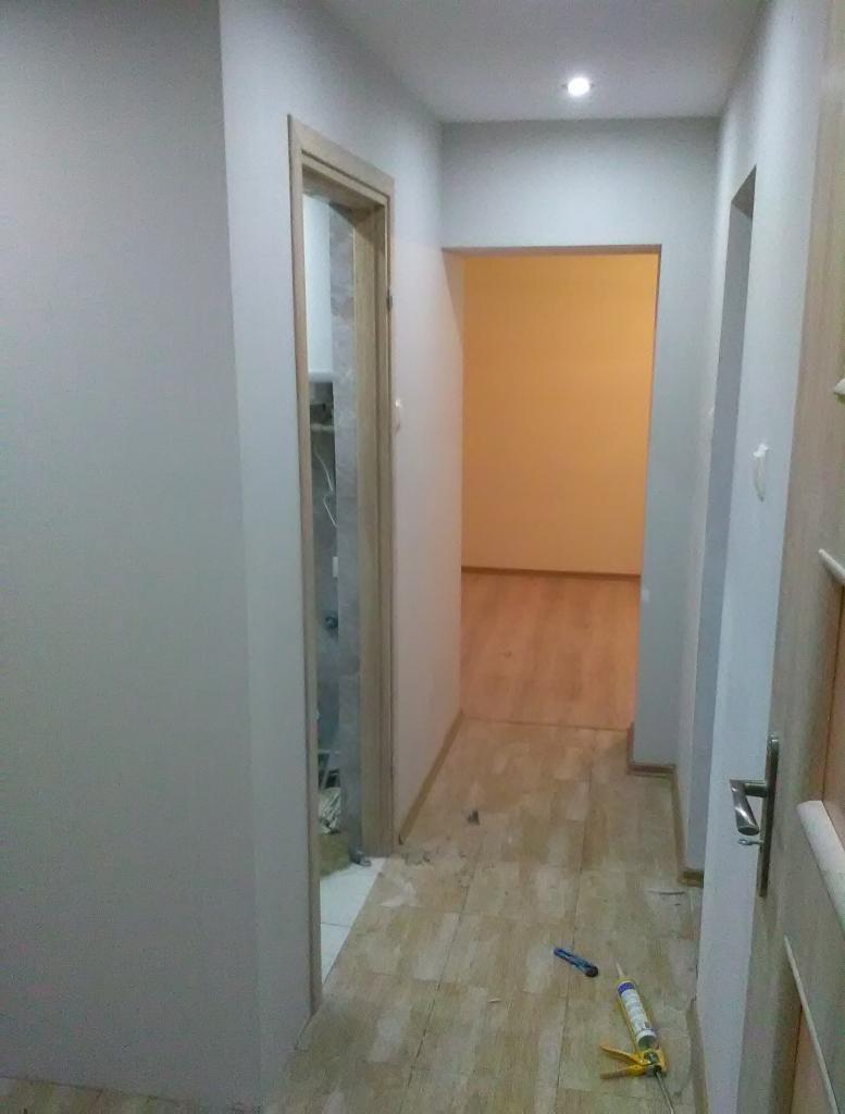 Korytarz w trakcie remontu: świeżo pomalowane ściany na szaro, częściowo ułożona podłoga z paneli, widoczny klej i nożyk do paneli.