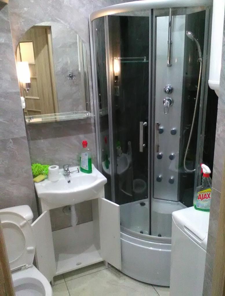 Mała łazienka z narożną kabiną prysznicową z hydromasażem, umywalką z szafką z otwartymi drzwiczkami i toaletą z otwartą klapą, widoczne środki czystości.