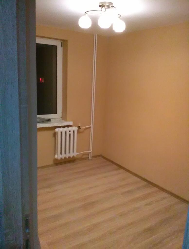 Pomalowane na brzoskwiniowo ściany pomieszczenia w Słubicach z drewnopodobnymi panelami podłogowymi, białym grzejnikiem pod oknem i lampą sufitową z trzema żarówkami.