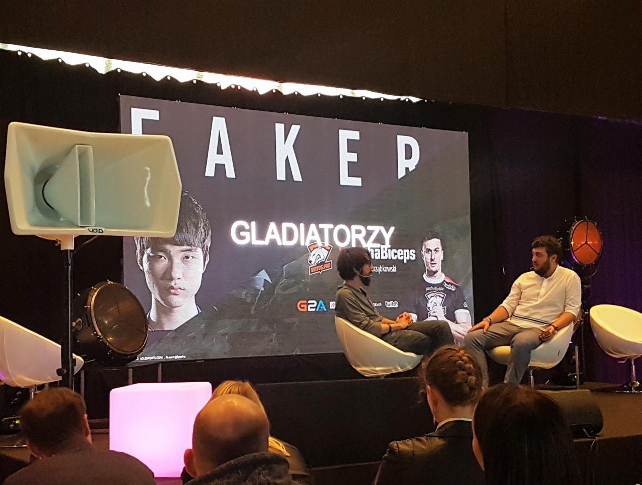 Konferencja z nagłośnieniem: scena z dwoma osobami siedzącymi na krzesłach, na tle ekranu z napisem 'FAKER' i wizerunkami graczy, z widocznymi głośnikami po lewej stronie i oświetleniem punktowym...