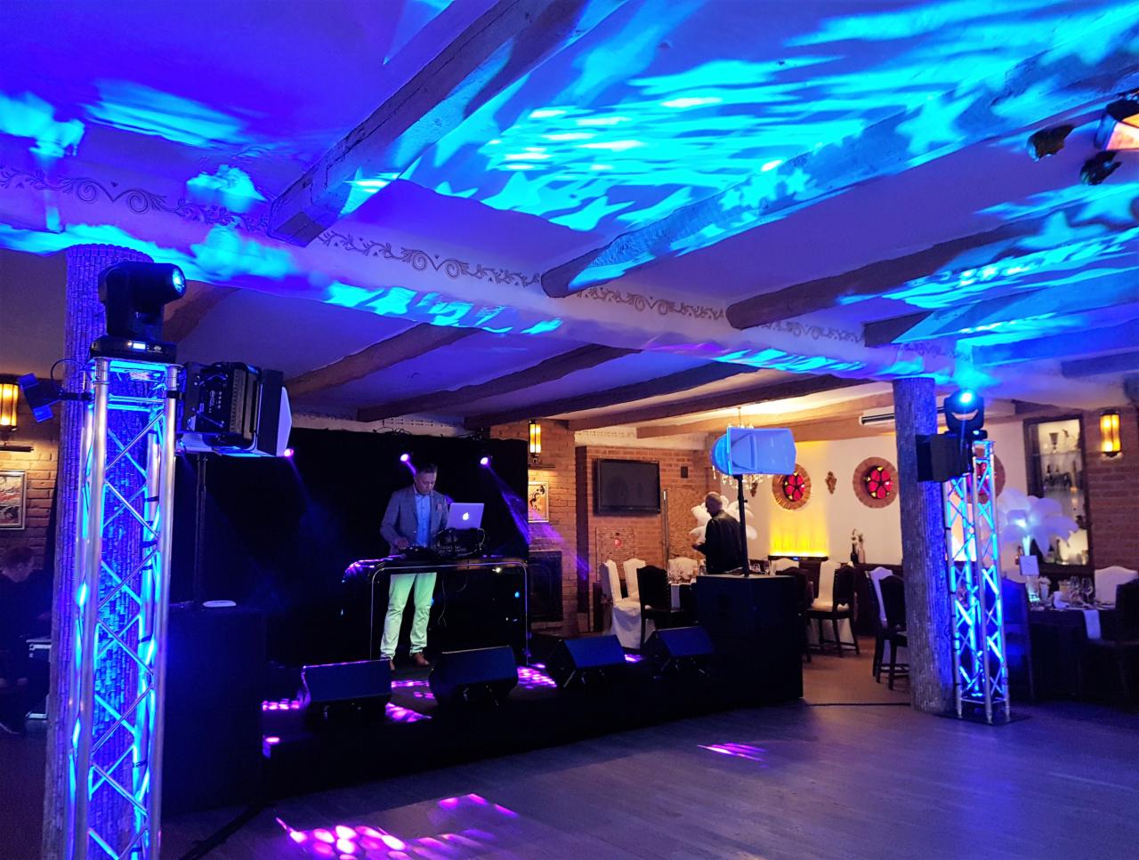 DJ pracujący na scenie z laptopem, oświetlenie sceniczne w odcieniach niebieskiego i fioletu, dekoracyjne oświetlenie sufitu w sali bankietowej.