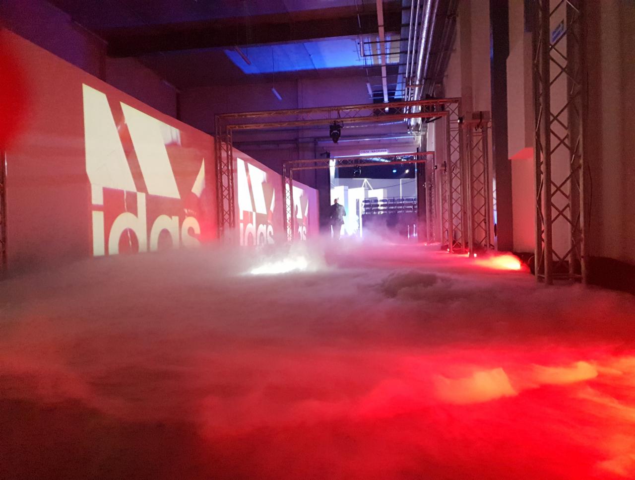 Scena eventowa z podświetlonym logo, efektami dymnymi i metalowymi konstrukcjami w tle, oświetlona czerwonym światłem.