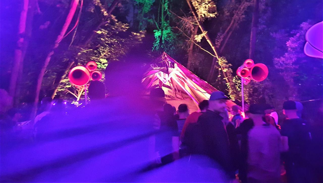 Scena plenerowa z namiotem oświetlonym na różowo i zielono, otoczonym przez drzewa i nagłośnienie w kształcie kwiatów, widzowie w tle.