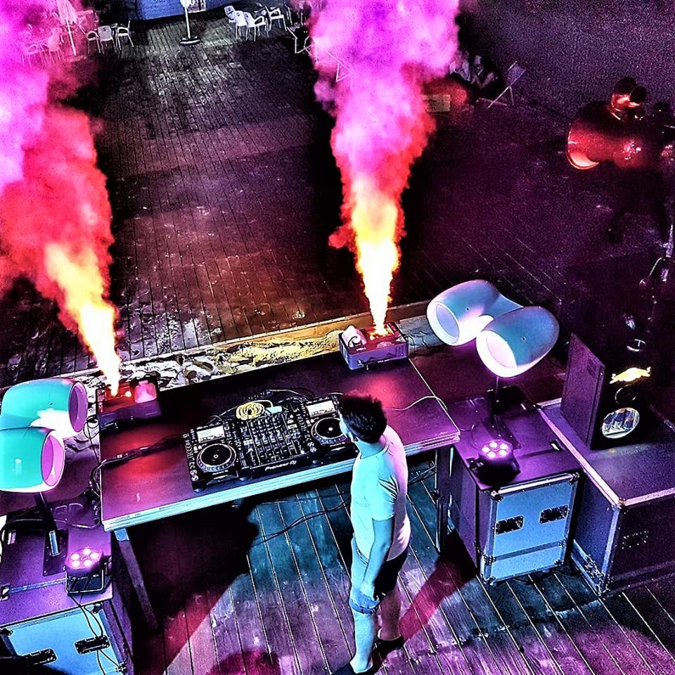 DJ stoi za konsoletą na drewnianym parkiecie, z tyłu dwa słupy różowego dymu i reflektory sceniczne podczas imprezy plenerowej.