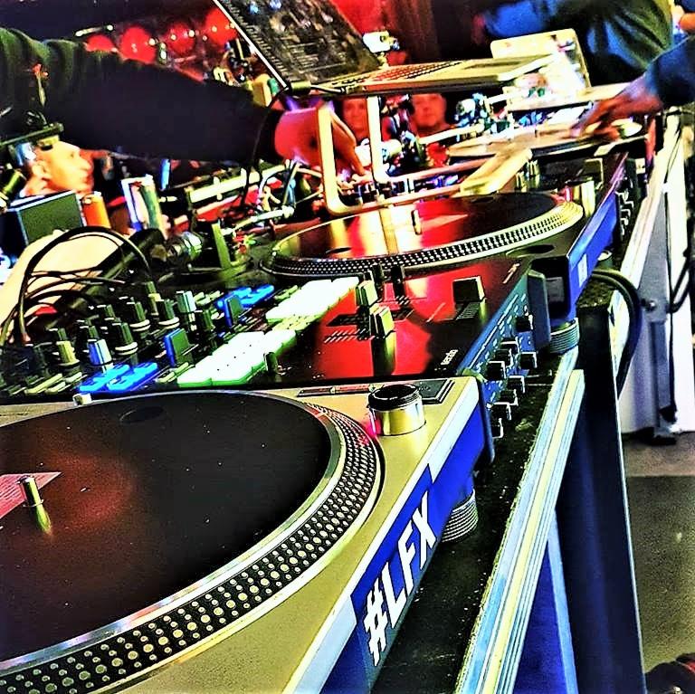 Zbliżenie na stanowisko DJ'a z dwoma gramofonami, mikserem i laptopem, oświetlone czerwonymi światłami, podczas występu na żywo.