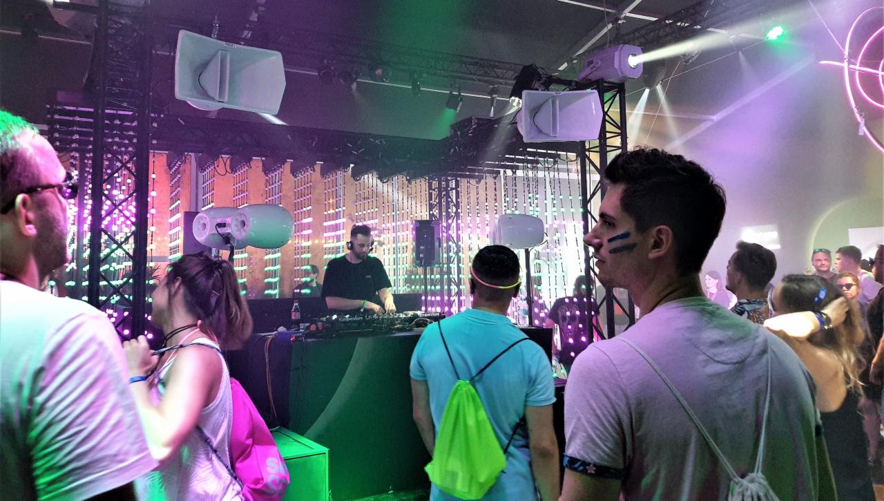 DJ za konsoletą podczas imprezy z oświetleniem scenicznym i nagłośnieniem, w tle dekoracja imitująca budynki, na pierwszym planie publiczność.