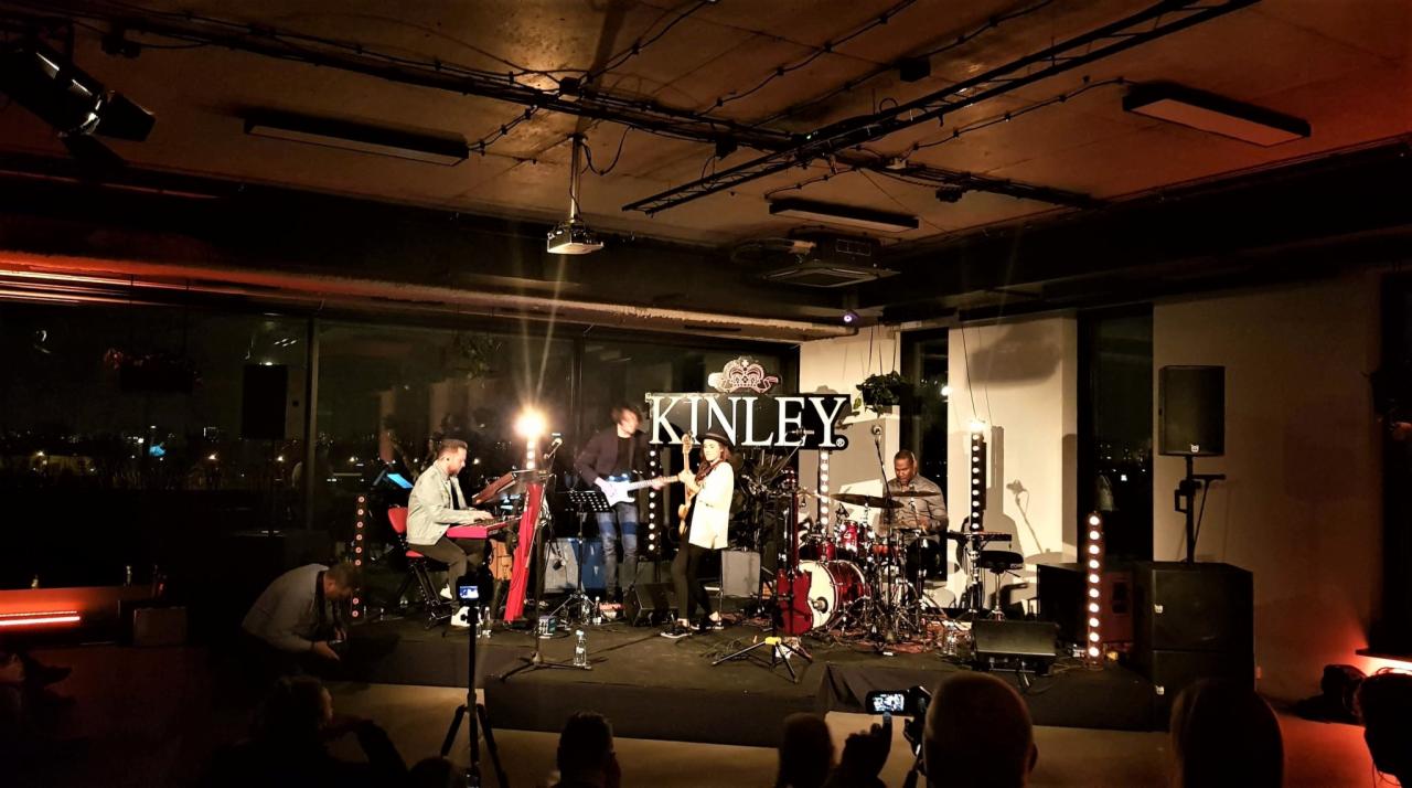 Zespół muzyczny Kinley na scenie podczas występu, widoczni muzycy grający na instrumentach, oświetlenie sceniczne i publiczność nagrywająca występ telefonami.