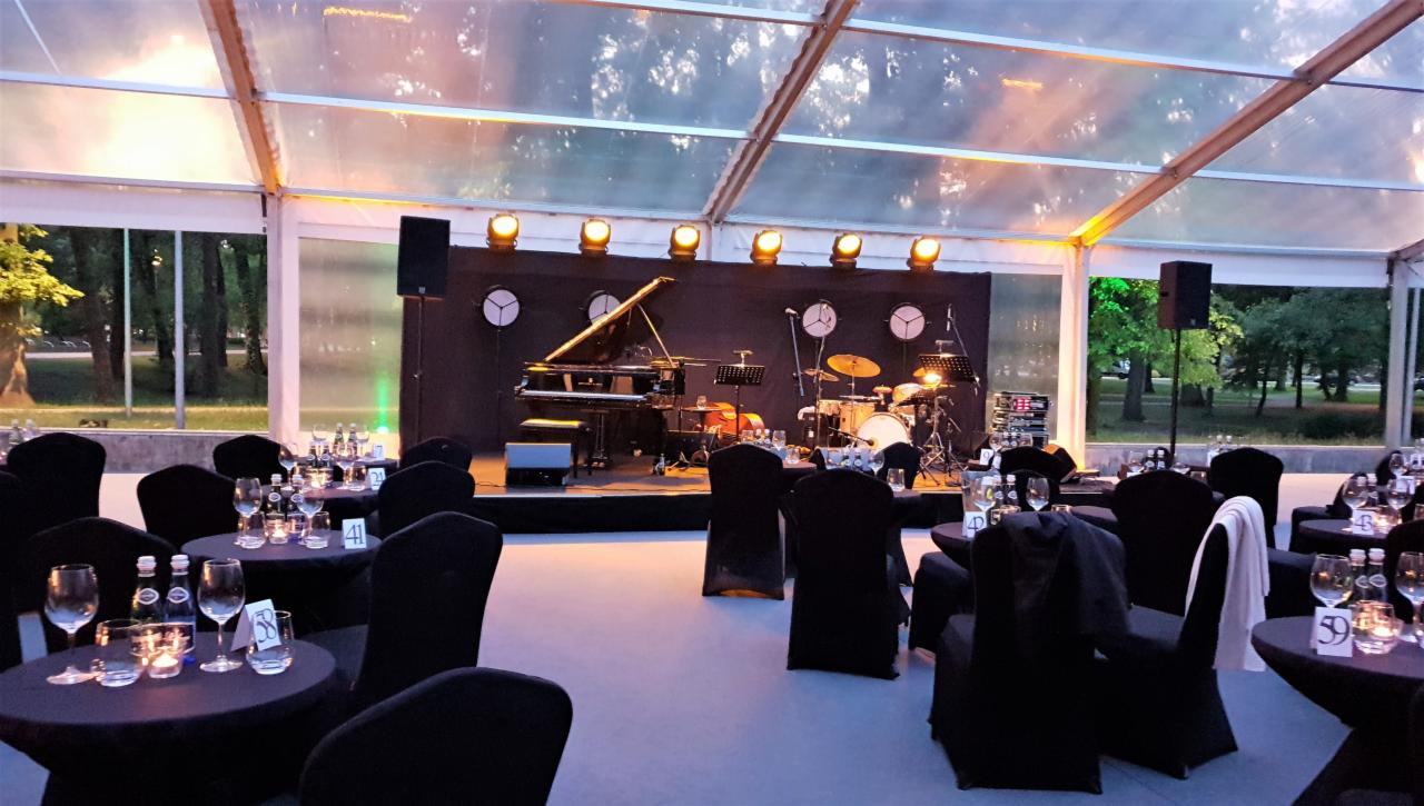 Scena z fortepianem i perkusją pod namiotem, przygotowana na imprezę z oświetleniem punktowym i stolikami dla gości.